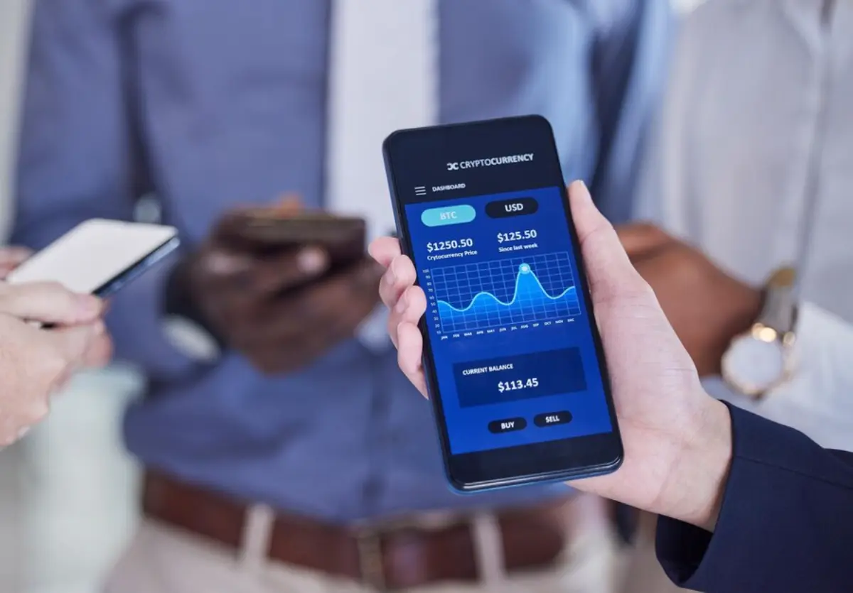 A person holding a smartphone displaying a cryptocurrency dashboard with price graphs, alongside others with mobile devices.