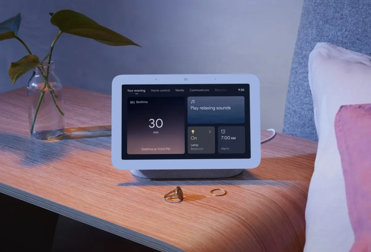 A smart display on a bedside table shows bedtime settings, with a plant and two rings beside it, indicating a cozy evening setup.