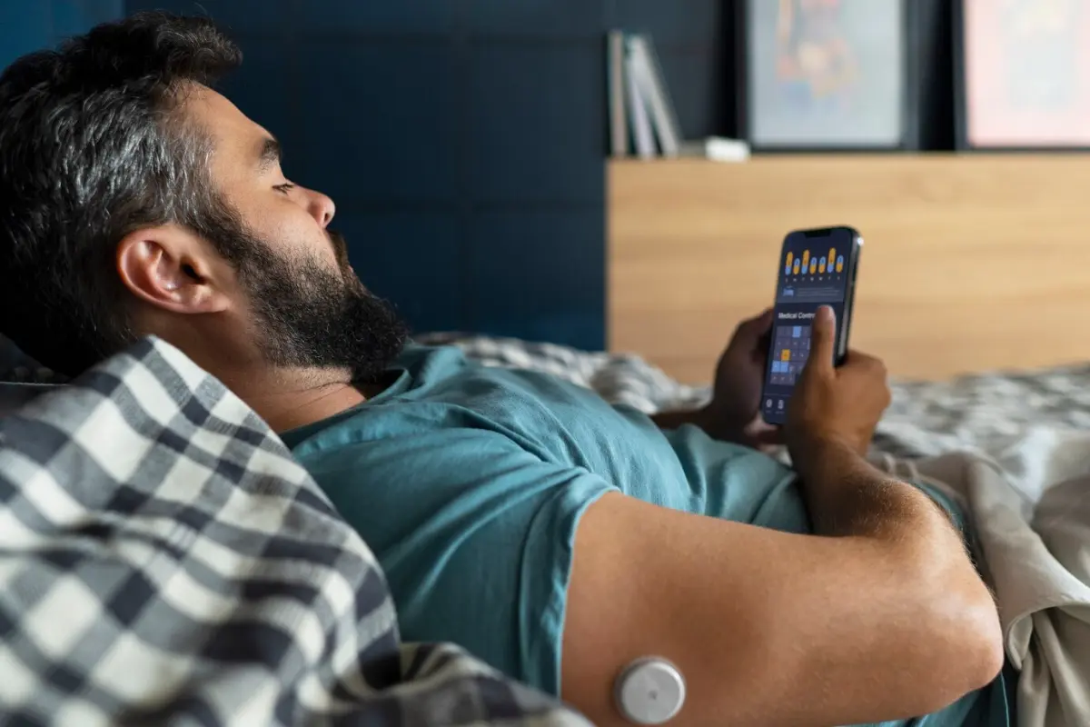 A person reclining in bed, wearing a blue shirt, using a smartphone to monitor health data, with a device attached to their arm.