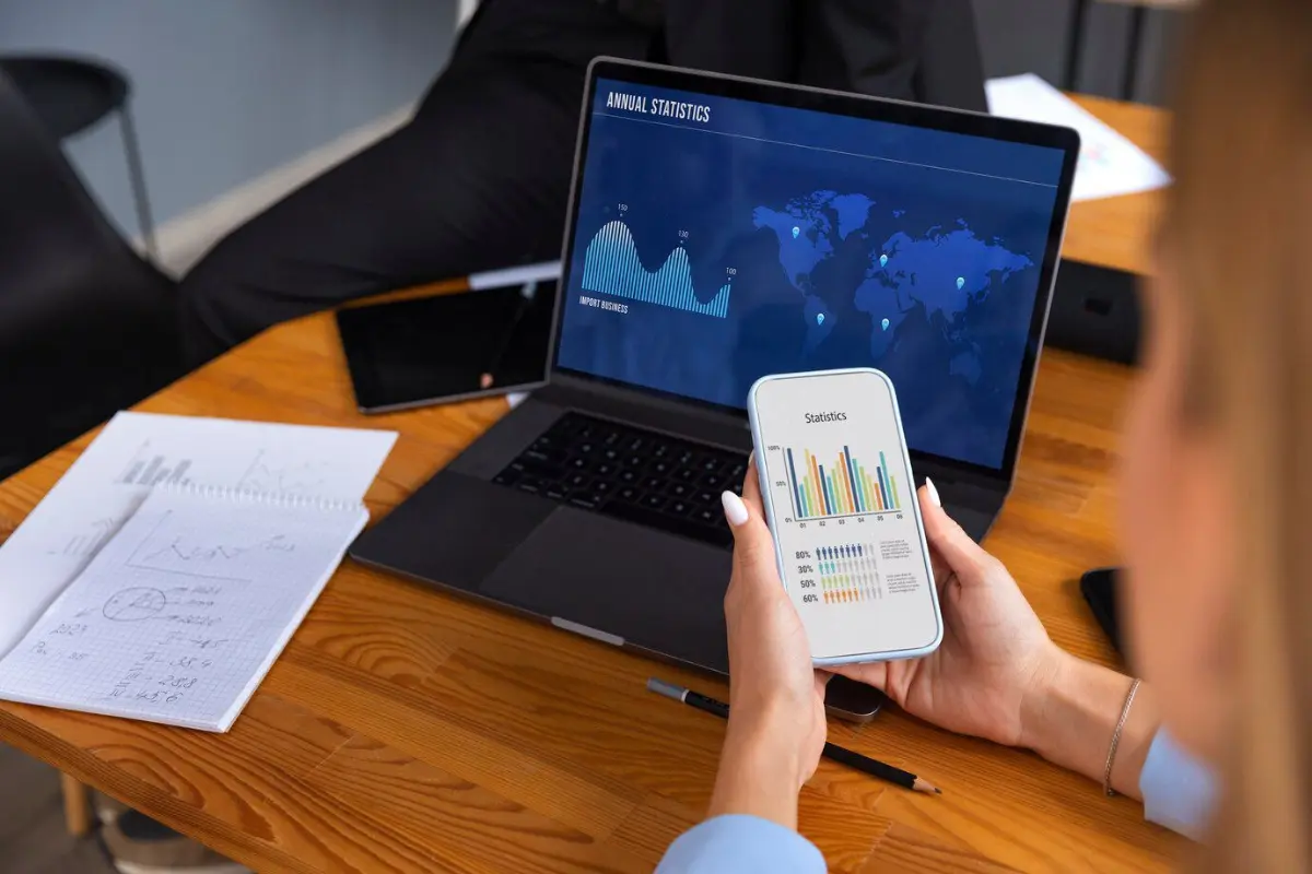 A person reviews annual statistics on a smartphone, while a laptop displays a world map and data graphs on a wooden desk.