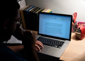 A person types on a laptop surrounded by books, stationery, and a colorful pencil sharpener on a wooden desk.