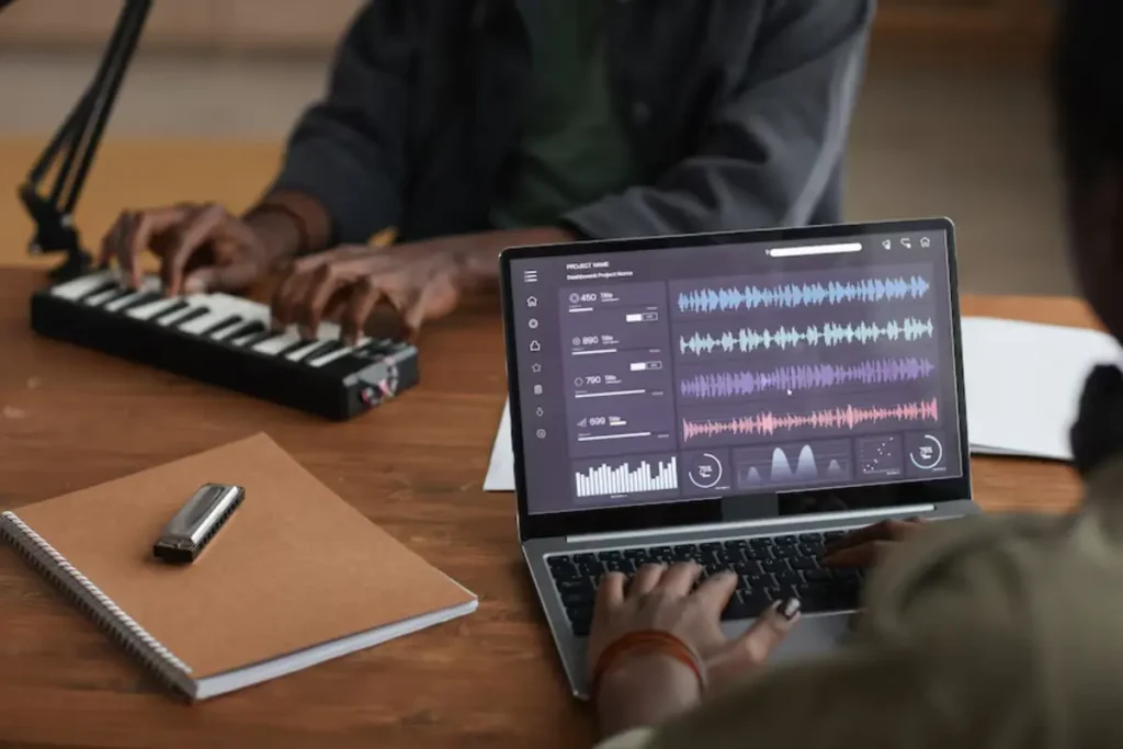 A person plays a mini keyboard while another edits audio on a laptop, showcasing a creative music production environment.