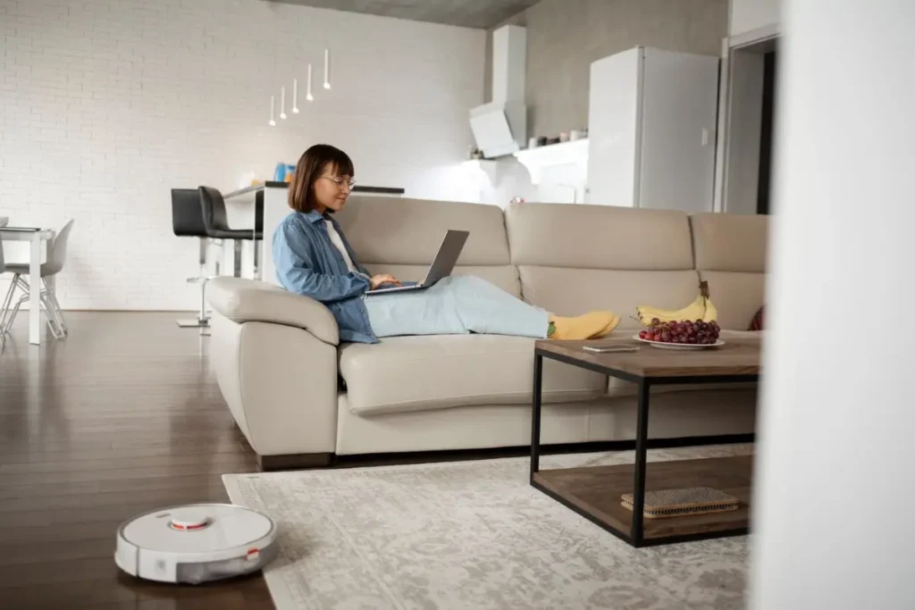 A woman sits on a beige sofa with a laptop, surrounded by fruit on a coffee table, while a robotic vacuum cleans the floor nearby.
