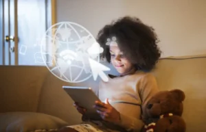A child sits on a couch, holding a tablet, surrounded by graphics of a globe and digital icons, with a teddy bear beside them.
