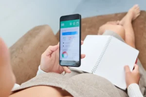 A pregnant woman holds a smartphone displaying a health app, while seated on a couch with an open notebook beside her.