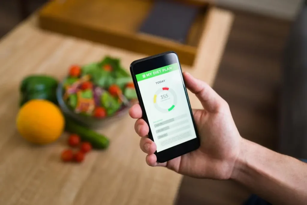 A hand holds a smartphone displaying a diet tracking app showing 355 calories consumed, with a bowl of salad and fresh vegetables nearby.